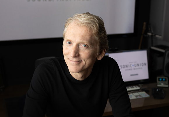 A smiling man with blond hair wearing a black long-sleeve shirt in a studio.