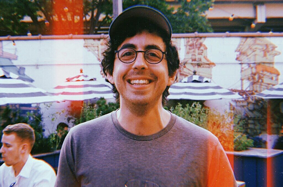 A smiling man with dark curly hair, glasses, and a dark cap wearing a grey t-shirt.