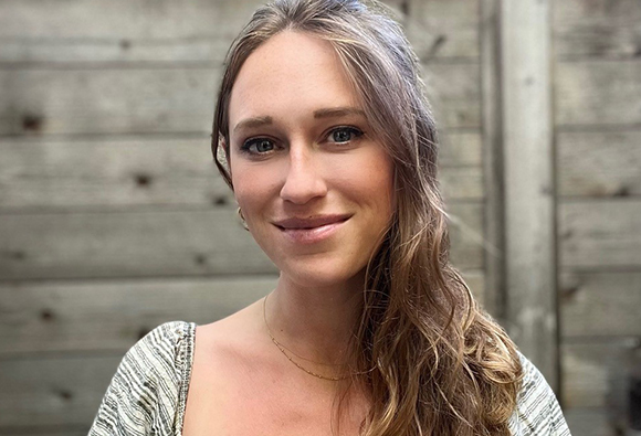 A woman with long wavy hair smiles toward the camera against a weathered wooden fence background.