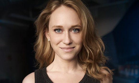 A blonde woman with blue eyes and a slight smile looks directly at the camera against a dark background.