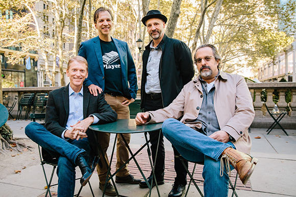 Four men standing and sitting around a small green table in a tree-lined outdoor courtyard.
