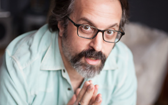 A man with dark hair, glasses, and a salt-and-pepper beard looks toward the camera with hands clasped.