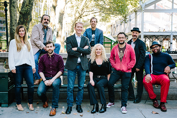 A group of nine diverse adults posing for a portrait in an outdoor urban park setting.