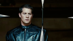 A man with short dark hair wearing a black leather jacket looks forward with a serious expression.
