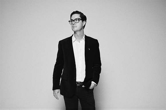 A man wearing glasses, a white shirt, and a dark blazer stands against a plain background.