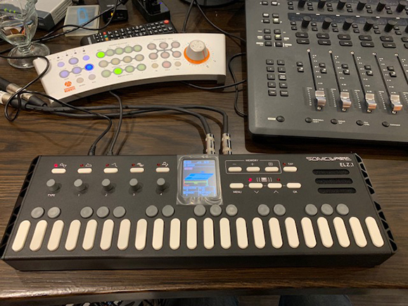 A black MIDI controller with white keys, a white control surface, and a large mixing console on a desk.