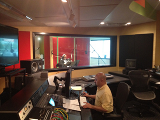 A man sits at a large audio mixing desk in a studio, looking toward a monitor.