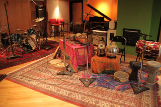 A music studio with drums, hand percussion on rugs, microphones, and a grand piano in the background.