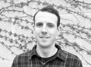 A man in a plaid shirt smiling in front of a wall covered in climbing vines.