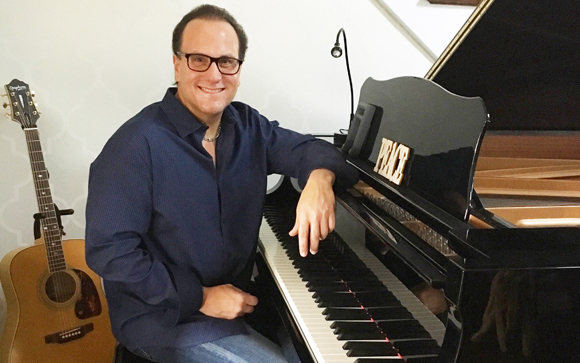 A man in a blue shirt sits at a black grand piano next to an acoustic guitar.