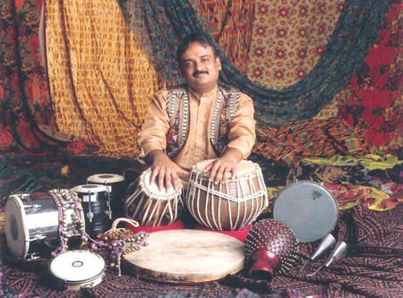 A man in traditional attire sits behind a pair of tabla drums surrounded by many other percussion instruments.