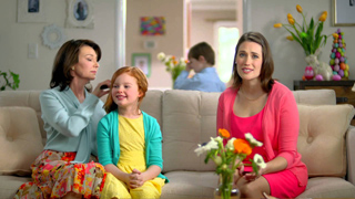 A woman in pink, a young girl in yellow, and a woman in light blue sit on a beige sofa indoors.