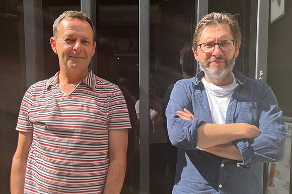 Two men stand in front of a dark building; one wears a striped polo, the other a blue shirt and glasses.
