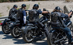 Four people in black leather vests and helmets sitting on motorcycles along a roadside.