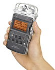 A person's hand holding a black and silver handheld digital audio recorder with a small screen.