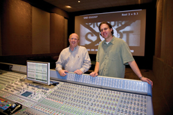 Two men stand behind a large professional audio mixing console in a dark studio.