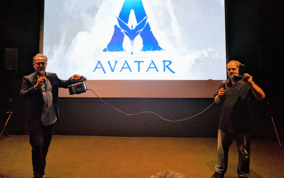 Two men stand on a stage holding a device connected by a long cable before a large Avatar movie screen.