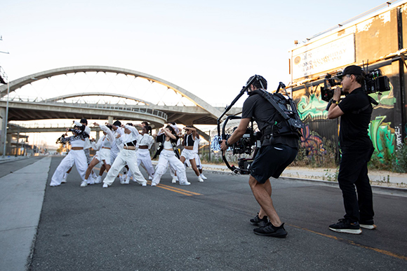 A camera operator with a stabilizer rig and a second cameraman film a group of dancers in white outfits on a street.