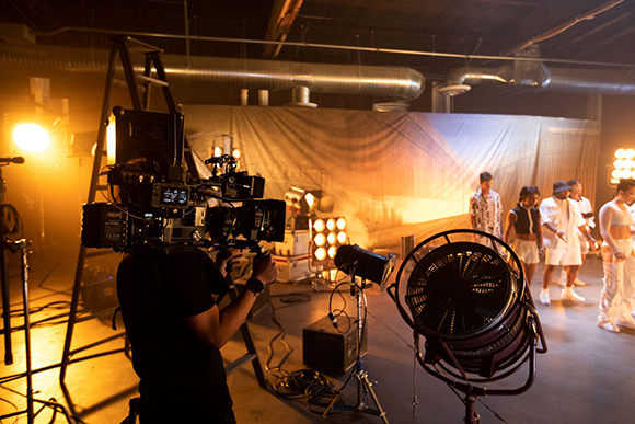 A camera operator films a group of people on a film set illuminated by warm studio lights.