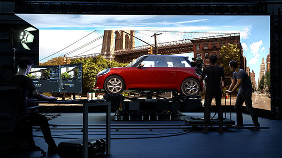 Film crew in a virtual production studio with a red car in front of a large LED screen showing a city bridge.
