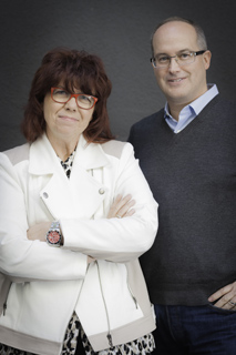 A woman with red hair and glasses in a white jacket stands next to a man in a grey sweater.
