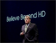 A man in a dark suit and patterned tie stands before a dark screen with the text 'Believe Beyond HD'.