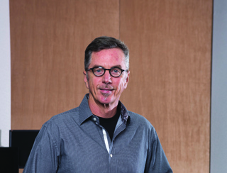 A man with short brown hair and glasses wearing a grey button-down shirt over a black shirt.