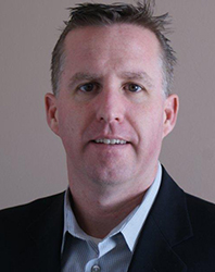 A man with short brown hair wearing a dark blazer and a light-colored button-down shirt.