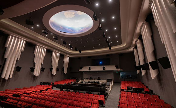 A wide view of a dark movie theater with rows of red seats and a large circular skylight-style ceiling light.