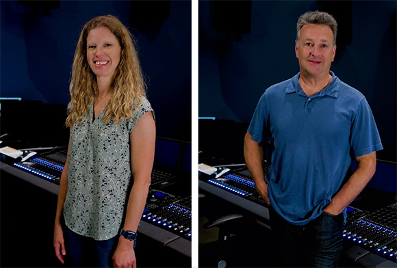 A woman in a patterned blouse and a man in a blue polo shirt stand in a dark recording studio.