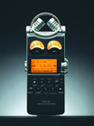 A black handheld digital audio recorder with two analog VU meters and an orange backlit LCD screen.