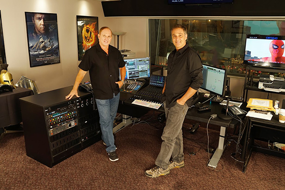 Two men standing in a studio filled with computer monitors, audio equipment, and movie posters.