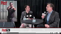 Two men in suits sit at a small round table with water bottles during a filmed interview.