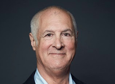 A headshot of an older man with thinning white hair and a slight smile against a dark background.