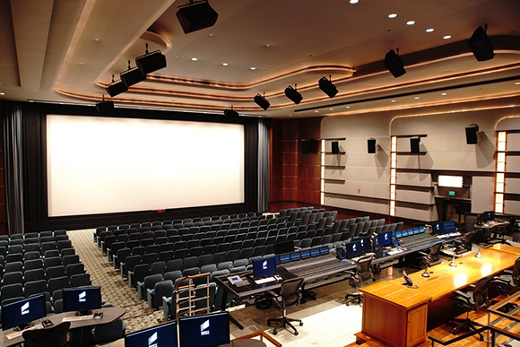 An empty auditorium featuring rows of dark blue seats facing a large, bright white projection screen.