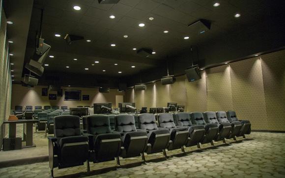 Rows of blue theater seats facing technical monitors and speakers in a darkened room.