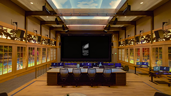 A large professional screening room with a central black screen, multiple monitors, and rows of desks.