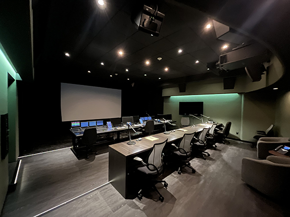 A dark media control room featuring a large projection screen, multiple computer monitors, and long desks with chairs.