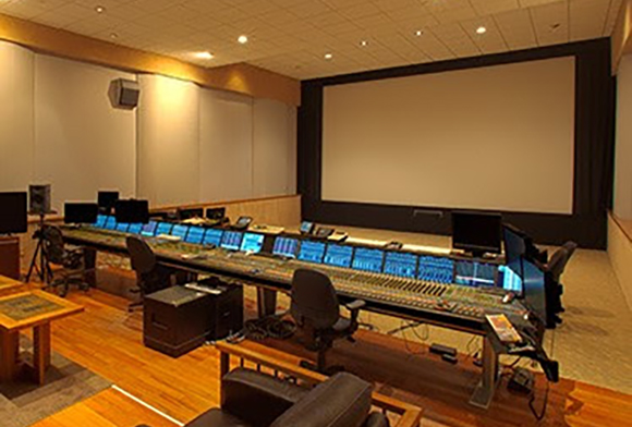 A large audio mixing console with multiple monitors in a studio with a large projection screen.