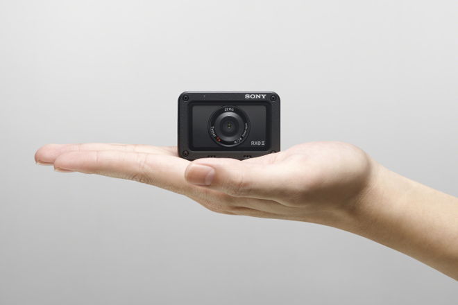 A person's open palm holding a small black Sony RX0 II compact camera against a light gray background.