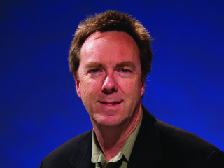 A man with short brown hair wearing a black jacket and olive shirt against a blue background.
