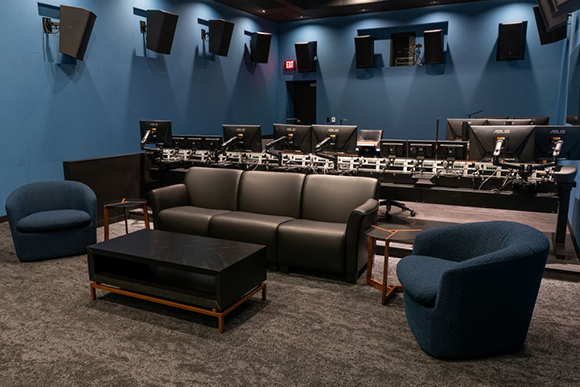A professional recording studio with blue walls, mixing consoles, large speakers, and a black leather sofa.