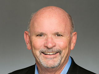 A smiling bald man with a grey goatee wearing a blue shirt and black blazer against a grey background.
