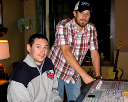 A man in a plaid shirt and cap stands by a mixing console while another man sits in front of it.