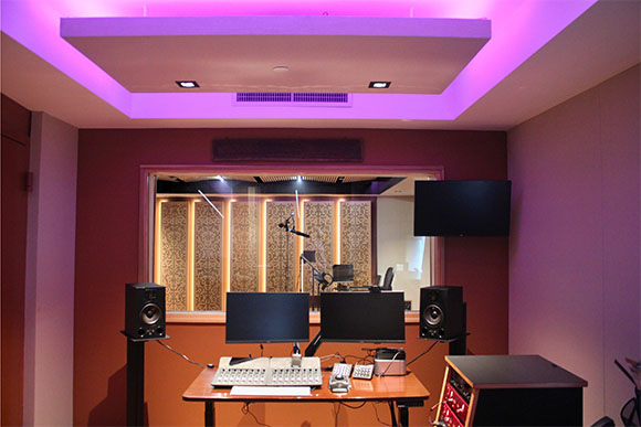 A recording studio control room with monitors, speakers, and a view into a soundproofed live room.