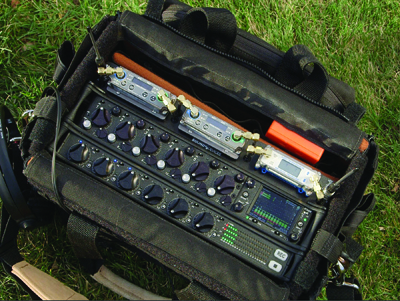 Audio mixer and multiple digital recorders inside an open black padded equipment bag on grass.