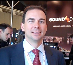 A man in a dark suit, light blue shirt, and red patterned tie smiling in front of a Sound Devices sign.
