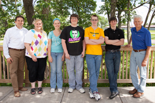 Six people of various ages stand in a line on a wooden deck in front of trees.