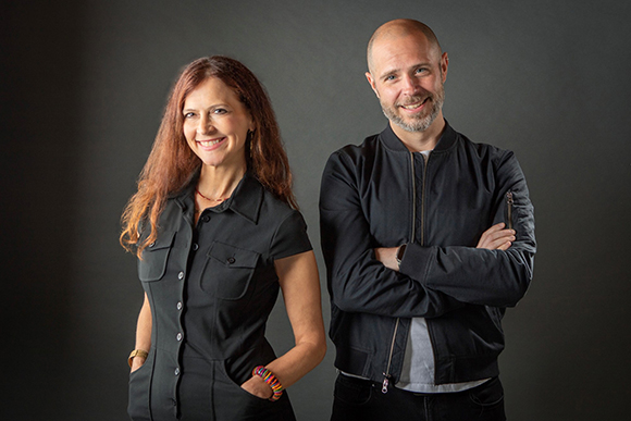 A smiling woman in a black button-down shirt and a man in a black bomber jacket standing side by side.