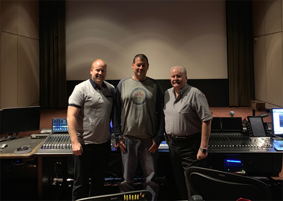 Three men standing together in a studio behind a large audio mixing console.